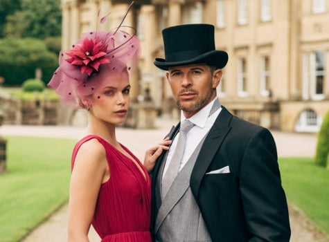 Formal portrait of a couple for a race day. The woman wears a bespoke magenta floral and netted fascinator with a rich raspberry dress. The man is in full morning dress with a black top hat and grey waistcoat, set against a classic English stately home.
