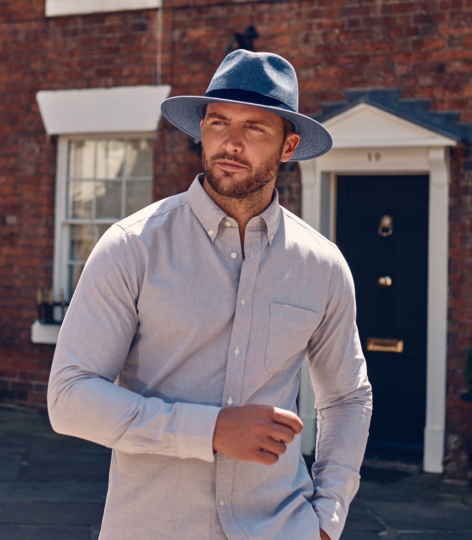 Man wearing a light blue shirt, with a darker blue straw fedora hat. Standing in front of a brick building.